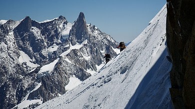 Two climbers ascending a steep snow-covered mountain slope with rugged peaks in the background during clear weather.