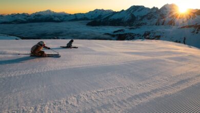 Powder snow groomed for skiing with two skiers on a mountain slope at sunrise, snow-capped peaks in the background, and a clear sky, highlighting winter sports excitement.