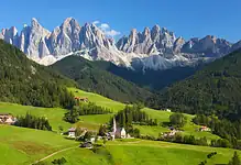 Vibrant green alpine landscape with a small village, church, and surrounding forests set against towering mountain peaks under a clear blue sky, perfect for winter and outdoor adventure enthusiasts.