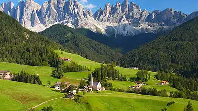 Vibrant green alpine landscape with a small village, church, and surrounding forests set against towering mountain peaks under a clear blue sky, perfect for winter and outdoor adventure enthusiasts.
