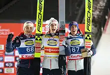 Three male cross-country skiers celebrating on podium after race, holding skis, with winter sports backdrop.