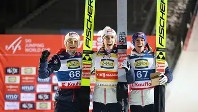 Three male cross-country skiers celebrating on podium after race, holding skis, with winter sports backdrop.