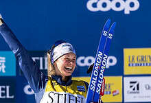 Excited female skier celebrating victory on podium at winter sports event with blue background and sponsor banners.