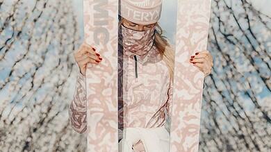 1. Person holding pink skis outdoors during spring with blossoming trees and blue sky, promoting winter sports gear and accessories for winter adventures.