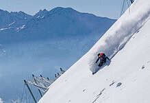 1. Brightly coloured skier in blue jacket and black pants carving down a snowy mountain slope with snow-capped peaks in the background, under a clear blue sky.