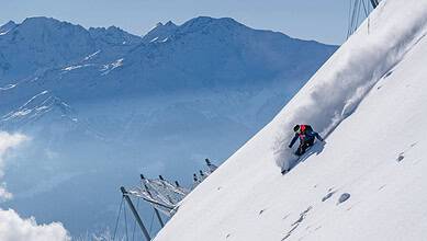 1. Brightly coloured skier in blue jacket and black pants carving down a snowy mountain slope with snow-capped peaks in the background, under a clear blue sky.