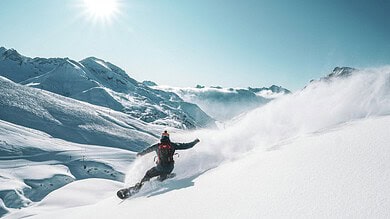 A snowboarder carving through fresh snow on a bright, sunny day in the mountains, with clear blue skies and snow-covered peaks in the background.
