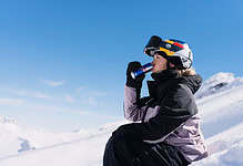 Female skier in winter gear drinking from a water bottle while sitting on snow-covered mountain slope, with clear blue sky and snowy peaks in the background.