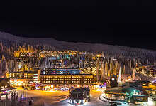 Nighttime view of a snowy mountain village illuminated with festive lights and decorated buildings. The scene captures the enchanting atmosphere of a winter holiday destination.