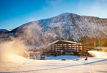A snow-covered ski resort with a wooden lodge and ski lifts against a backdrop of mountains under a clear blue sky.