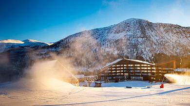 A snow-covered ski resort with a wooden lodge and ski lifts against a backdrop of mountains under a clear blue sky.