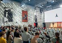 Large audience attending a conference with a speaker on stage and large screens in a modern event hall.