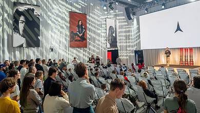 Large audience attending a conference with a speaker on stage and large screens in a modern event hall.