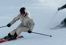 A skier in white winter gear and a black helmet skiing down fresh snow with a companion in the background, enjoying a winter day.