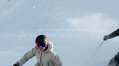 A skier in white winter gear and a black helmet skiing down fresh snow with a companion in the background, enjoying a winter day.