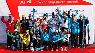 Group of winter sports athletes celebrating on the podium with medals and skis, in front of a red backdrop with Audi branding.