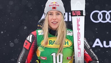 Female skier in racing gear with bib number 10, snowy backdrop, Red Bull beanie, holding a ski pole, promotional branding visible.