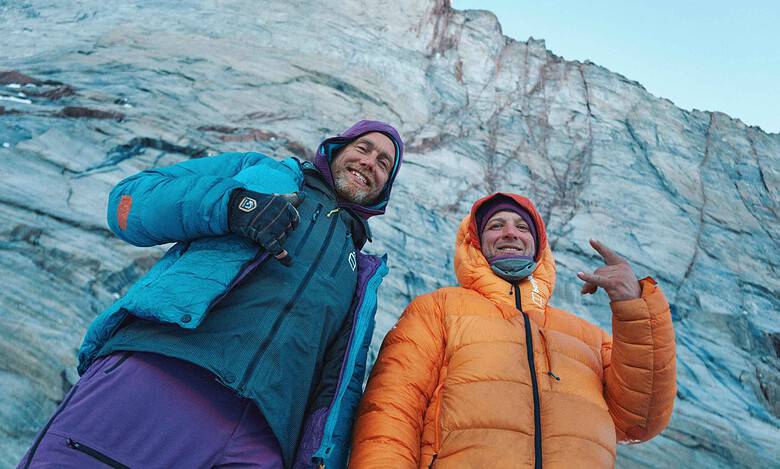 Two explorers in colorful winter jackets standing in front of a glacier, smiling and enjoying the winter outdoor adventure.