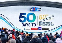 Crowd watching a luge race at the Eberspächer Luge World Cup with a large banner displaying '50 Days To' and event logos.