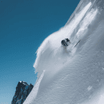 Skier descending a steep, snow-covered mountain slope during bright winter day, showcasing thrilling winter sports activity.