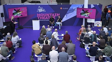 Audience watching a panel discussion at the mountain theatre during a winter sports event, with screens displaying promotional content and a scenic mountain backdrop.
