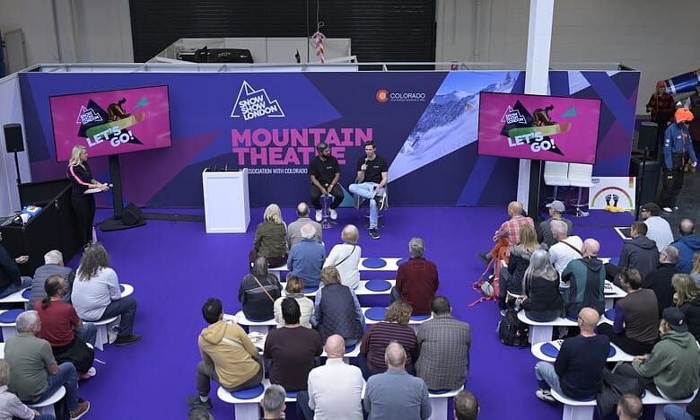 Audience watching a panel discussion at the mountain theatre during a winter sports event, with screens displaying promotional content and a scenic mountain backdrop.