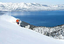 Skier descending a snowy mountain slope with a stunning lake and snow-covered mountains in the background, capturing the thrill of winter sports.