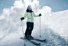 Skier in pale yellow jacket and black pants navigating fresh powder on a sunny snowy slope with blue sky.